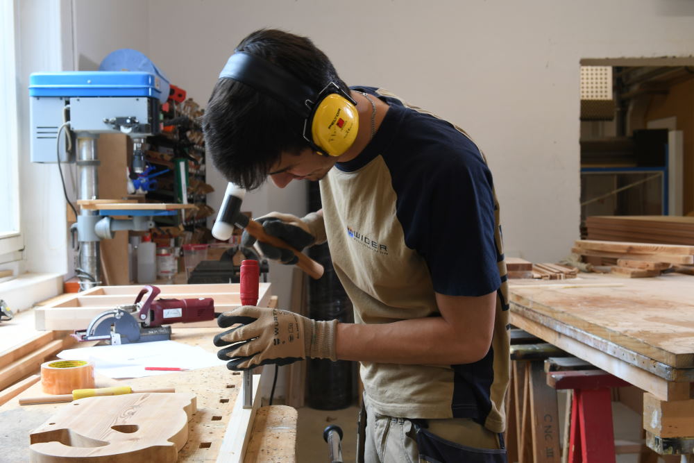 Ein Schreiner mit Lärmschutz-Kopfhörer bearbeitet Holz in einer Werkstatt.