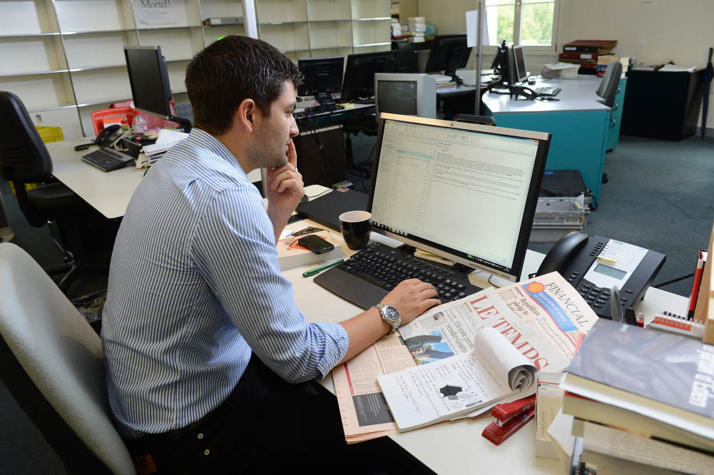 Ein Mann steht vor dem Bildschirm und auf seinem Schreibtisch liegen Zeitungen.