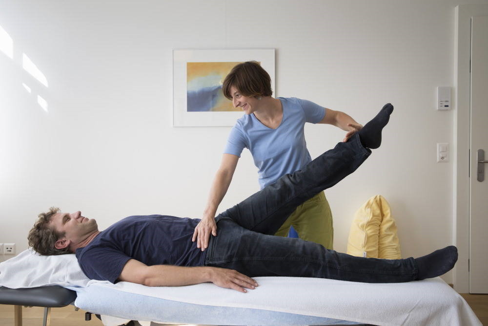 Eine Fachfrau hebt während einer physiotherapeutischen Übung das Bein eines Patienten an.