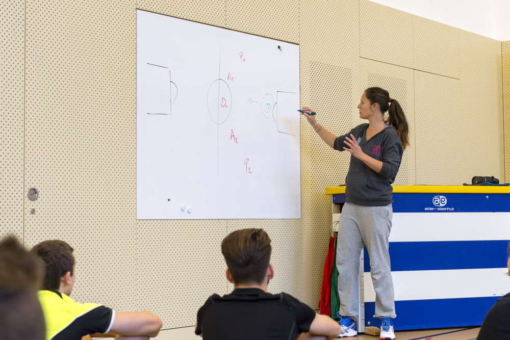 Eine Lehrperson erklärt den Schülerinnen und Schülern die Spielregeln auf einem Whiteboard in der Turnhalle.