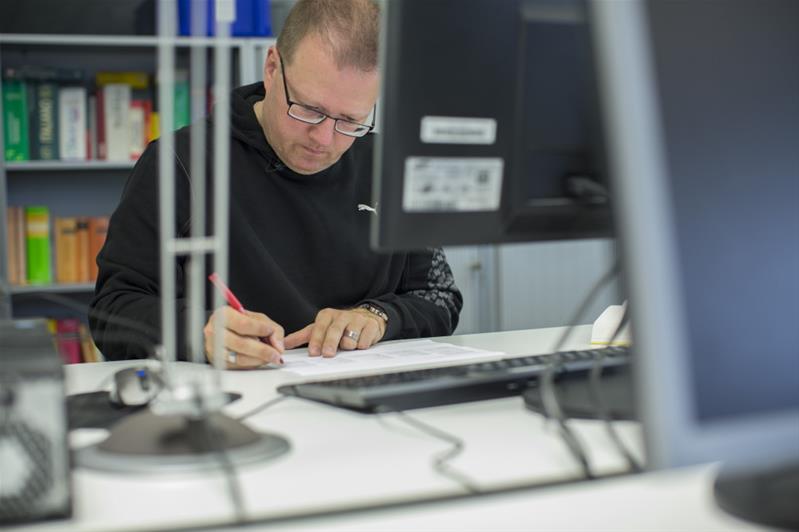 Ein Mann vor einem Bildschirm korrigiert ein gedrucktes Dokument mit einem roten Stift.