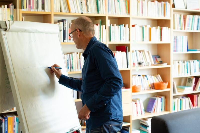 Ein Berater zeichnet etwas auf einem Flipchart auf, im Hintergrund stehen an der Wand mehrere Bücherregale.