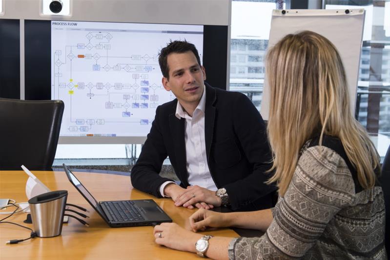 Ein Manager diskutiert mit einer Frau in einem Sitzungsraum. Ein Schema ist auf dem Bildschirm im Hintergrund sichtbar.